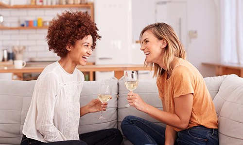 Two women sitting on a couch drinking wine and laughing.