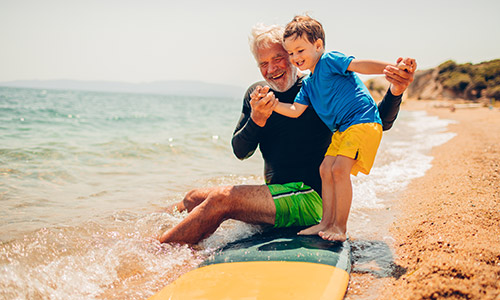 Man holding a little boy on a surfboard on the beach sand.